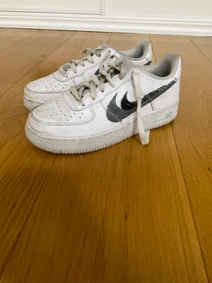 Nike Air Force 1 vita sneakers - Nike Air Force 1 sneakers i vitt skinn med klassisk svart swoosh på sidan. Låg modell med rund tå, snörning och platt sula. Perforerade detaljer på tån för extra ventilation. Ikonisk Nike Air-logga på hälen. Perfekt för dig som gillar streetstyle.