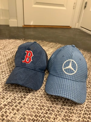 Två blå kepsar: Boston Red Sox & Mercedes - Säljer två snygga kepsar! En mörkblå Boston Red Sox-keps med röd B-logga framtill och en ljusblå Mercedes-keps med vitt mönster och stor logga. Båda har böjd skärm och justerbar rem bak. Perfekt för dig som gillar sport eller bilar.