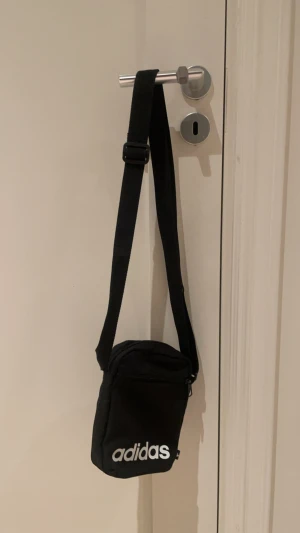 Adidas axelväska  - Snygg svart axelväska från Adidas med justerbar rem och vit logga framtill. Väskan har ett huvudfack med dragkedja och är tillverkad i slitstark polyester. Perfekt storlek för mobil, plånbok och andra småsaker. Väldigt fin och som ny.