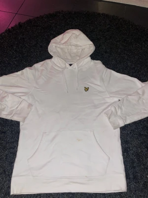 Vit hoodie från Lyle & Scott i S - Vit hoodie från Lyle & Scott med klassisk gul broderad logga på bröstet. Tröjan har huva med snörning och en stor magficka. Tillverkad i mjuk bomull, perfekt för chill dagar. Långärmad och med ribbade muddar för extra komfort.