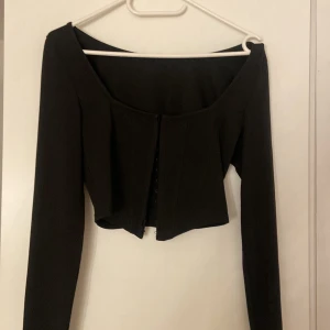 Svart croppad korsett-topp - Säljer en svart croppad korsett-topp med lång ärm och snygg offshoulder-skärning. Toppen har hakar framtill och markerade sömmar som ger en cool siluett. Perfekt för dig som gillar trendiga och stilrena plagg med edge. Funkar som xs också 