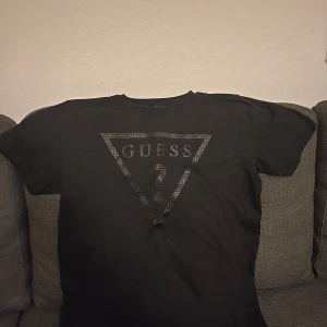 Svart t-shirt från Guess Kids - Svart t-shirt från Guess Kids med klassisk triangellogga och frågetecken i glansigt tryck på bröstet. Modellen är kortärmad och har rund hals. Materialet är mjuk bomull som känns skön mot huden. Perfekt för dig som gillar stilrena och ikoniska märken. Sitter som en liten Small