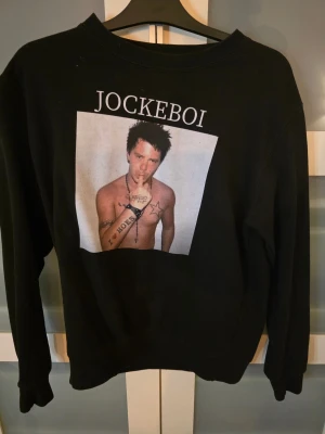 Svart sweatshirt Jockeboi JustHoods M - Svart sweatshirt från JustHoods i storlek M med tryck både fram och bak. Framsidan har en stor bild med texten 'JOCKEBOI' och baksidan har tryck med texten 'JOJO BITCHES'. Tröjan har rund hals och långa ärmar, perfekt för dig som gillar unika statement-plagg.