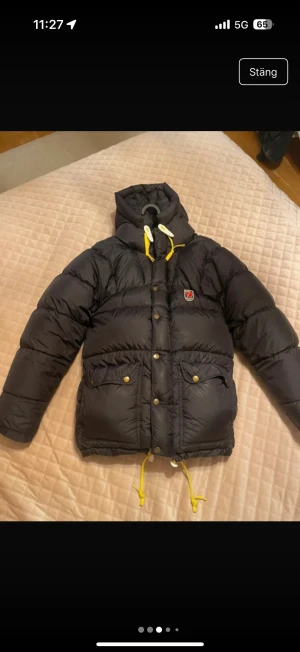 Svart dunjacka från Fjällräven  - Svart dunjacka från Fjällräven med gul snörning i huva och nederkant, samt tryckknappar framtill. Jackan har två stora fickor med lock och Fjällräven-logga på bröstet. Puffig modell med huva och lång ärm, perfekt för kalla dagar. Lånade bilder !😁