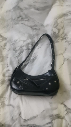 Svart axelväska med nitar och dragkedja - Cool svart axelväska i skinnimitation med silvriga nitar och detaljer. Väskan har en snygg böjd form, en framficka med dragkedja och justerbar axelrem med metallhål. Perfekt för att ge din outfit lite edge. Pris kan diskuteras 💞