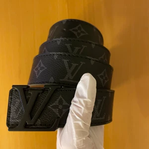 Svart skärp med monogram - Säljer ett svart bälte i skinn med ikoniskt monogrammönster och ett -spänne i mörk metall. Bältet har en klassisk och stilren look som lyfter vilken outfit som helst. Kommer med originalask och kvitto.