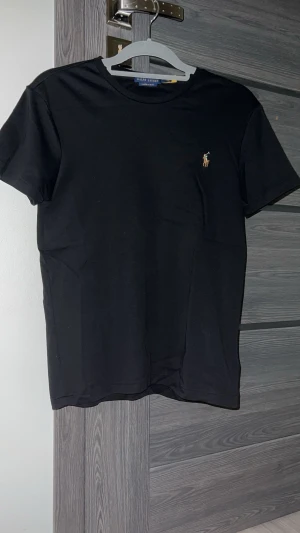 Svart t-shirt från Polo Ralph Lauren - Svart t-shirt från Polo Ralph Lauren med klassisk rund hals och liten broderad logga på bröstet. Modellen är custom slim fit och har korta ärmar. Materialet är mjuk bomull som känns skön mot huden.