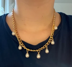 Guldhalsband med pärldroppar - Snyggt halsband i guldtonad kedja med sju hängande droppformade pärlor. Kedjan är grov och ger ett lyxigt intryck, medan pärlorna ger en elegant touch. Perfekt för dig som gillar att sticka ut med accessoarer som syns. Handgjort
