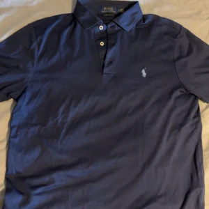 Marinblå pikétröja från Ralph Lauren - Snygg marinblå pikétröja från Ralph Lauren. Passformen är custom slim fit så sitter riktigt bra på. Skicket är som nytt, finns inga skador eller fläckar 
