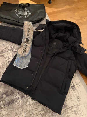 Svart dunjacka från Canada Goose L - Säljer en svart dunjacka från Canada Goose med huva och avtagbar pälskant. Jackan har en klassisk patch på ärmen, dragkedja och tryckknappar framtill. Perfekt för kalla vinterdagar och har en riktigt schysst puffad look. Skick 9,5/10 , Helt fräsch. Kan tänka mig byta mot en annan jacka eller något annat beroende på vad.Den är i storlek L men passar folk i storlek M också. Skriv bara för mer info