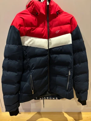 Pufferjacka från Five Seasons - Snygg pufferjacka från Five Seasons med färgblock i rött, vitt och mörkblått. Jackan har huva, två dragkedjefickor och logga på bröstet. Perfekt för kalla dagar med sin tjocka vaddering och sportiga look. Designad i Sverige, med coolt tryck på insidan. Parfekt till vintern 