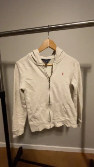 Beige hoodie från Polo Ralph Lauren  - Säljer en vit hoodie från Polo Ralph Lauren i storlek XL. Tröjan har huva, dragkedja framtill och klassisk röd broderad logga på bröstet. Tillverkad i mjuk bomull med ribbade muddar och fickor framtill. Perfekt för chill dagar.