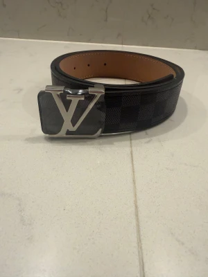 Louis Vuitton rutig skinnbälte svart - Säljer ett Louis Vuitton bälte i svart och grått med klassiskt rutigt Damier-mönster. Bältet har en stor silvrig LV-spänne och är tillverkat i äkta skinn. Insidan är beige och bältet är tillverkat i Frankrike. Perfekt för dig som vill ha en lyxig och stilren accessoar.