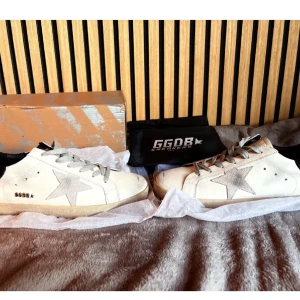 Golden Goose Superstar sneakers - Golden Goose Superstar sneakers i vitt skinn med grå mockastjärna på sidan och svart hälparti. Klassisk låg modell med grå snörning och beige sula. Snygg och trendig design, perfekt för dig som gillar streetstyle och italiensk kvalitet.