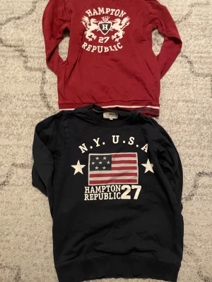 Två långärmade tröjor Hampton Republic - Säljer två snygga långärmade tröjor från Hampton Republic. En vinröd med vitt tryck och coolt emblem, samt en mörkblå med amerikansk flagga och stjärnor. Båda har rund hals och tryck framtill, perfekta för dig som gillar preppy stil.