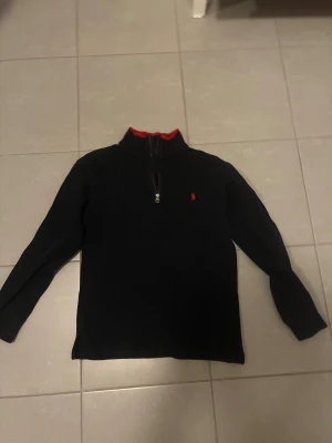 Svart half zip tröja från Polo Ralph Lauren - Svart långärmad tröja från Polo Ralph Lauren med röd broderad logga på bröstet och röd insida vid kragen. Tröjan har half zip-dragkedja och är gjord i 100% bomull, perfekt för dig som gillar klassisk stil med en sportig touch.