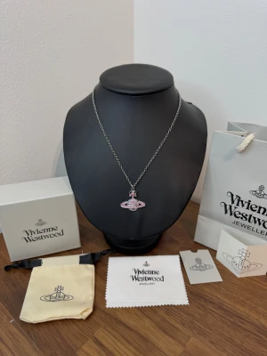 Vivienne Westwood halsband med rosa stenar - Unikt halsband från Vivienne Westwood med ikonisk orb-berlock dekorerad med glittrande rosa stenar. Kedjan är i silverfärgad metall och har en liten tag med loggan. Perfekt statement piece för dig som gillar att sticka ut. Kommer med: gift bag, gift box, dust cloth, care instructions, dust bag (som på bilden).