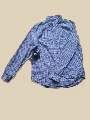 Blårutig skjorta från Ralph Lauren - Snygg blårutig skjorta från Ralph Lauren med klassisk krage och knappar hela vägen. Skjortan har lång ärm och det ikoniska röda Polo-logot på bröstet. Perfekt för dig som gillar stilrena och tidlösa plagg med en ungdomlig touch.