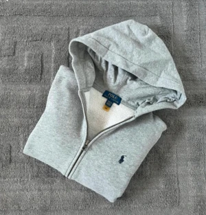 Grå hoodie från Polo Ralph Lauren - Snygg grå hoodie från Polo Ralph Lauren med dragkedja och klassisk broderad logga på bröstet. Tröjan har huva, kängurufickor och är tillverkad i mjuk bomullsmix. Perfekt för chill dagar eller när du vill ha en clean och enkel look.