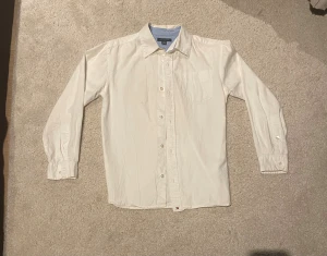 Vit skjorta från Tommy Hilfiger - Vit skjorta från Tommy Hilfiger med klassisk krage och ljusblå insida vid halsen. Skjortan har långa ärmar och knappar med logotyp. Liten broderad Hilfiger-flagga vid nederkanten. Perfekt för dig som gillar stilrena och enkla plagg.