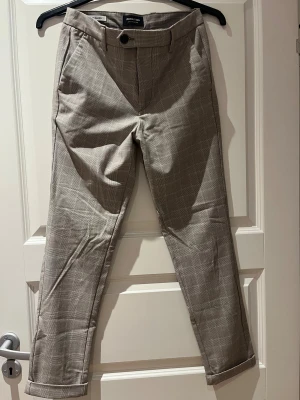 Rutig kostymbyxa Jack & Jones Slim Marco - Snygga rutiga kostymbyxor från Jack & Jones i modellen Slim Marco. Byxorna är beige med diskret rutmönster, har slim passform, bälteshällor, framfickor och bakfickor med knappar. Perfekta för en stilren look.