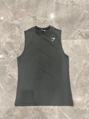 Mörkgrå ärmlös linnetopp från Gymshark - Säljer en stilren mörkgrå linnetopp från Gymshark, perfekt för träning. Toppen är ärmlös med rund hals och har ett vitt Gymshark-logotryck på bröstet. Materialet är lätt och ventilerande polyester, vilket gör den skön att bära under intensiva pass.