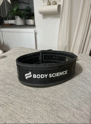 Svart träningsbälte Body Science - Stabilt svart träningsbälte från Body Science med vit kontrastsöm och metallspänne. Bältet är brett och rejält, perfekt för gymmet och tunga lyft. Snyggt tryck med logga framtill och robust konstruktion i skinn för extra stöd vid träning.