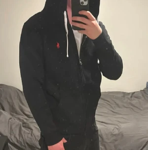 Svart hoodie från Polo Ralph Lauren - Svart hoodie från Polo Ralph Lauren med dragkedja och vit snörning i huvan. Klassisk röd logga broderad på bröstet. Tröjan har långärmade ärmar och är gjord i mjuk bomull, perfekt för chill dagar. Snygg och enkel stil som funkar till jeans eller joggers.