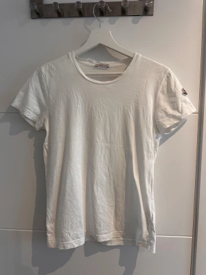 Vit Moncler t-shirt i bomull - Klassisk vit t-shirt från Moncler i mjuk bomull. T-shirten har rund halsringning, korta ärmar och Monclers ikoniska logga på vänster ärm. Enkel och clean design som funkar till allt. Perfekt basplagg för dig som gillar stilrena märkeskläder.