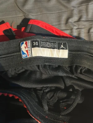 Chicago Bulls basketshorts Jordan NBA - Säljer ett par svarta och röda Chicago Bulls basketshorts från Jordan NBA Authentics. Shortsen har elastisk midja med snörning, Bulls-logga på benen och vita Jordan-loggan. Snygga detaljer med stjärnor och kontrastpaneler. Perfekta för basket eller streetwear.