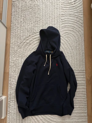 Blå hoodie från Polo Ralph Lauren - Snygg marinblå hoodie från Polo Ralph Lauren med klassisk röd logobrodyr på bröstet. Perfekt för dig som gillar stilren och enkel design med premiumkänsla. Tröjan är knappt använd och fick den som present, är för liten på mig. Brukar vanligtvis ha L.