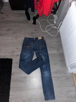 Mörkblå jeans från Nudie Jeans - Snygga mörkblå jeans från Nudie Jeans med klassisk femficksdesign och kontrastsömmar. Jeansen har en rak passform och är tillverkade i slitstarkt denim med subtila tvättade detaljer. Perfekta för dig som gillar stilrena och tidlösa jeans. 32/34