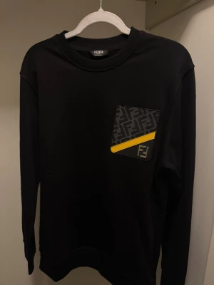 Svart Fendi sweatshirt med fickdetalj - Svart sweatshirt från Fendi med lång ärm och rund hals. På bröstet finns en snygg ficka med Fendi-logga, mönstrad i grått och svart samt en gul diagonal detalj. Perfekt för dig som gillar exklusiva streetwear-plagg med unik design.