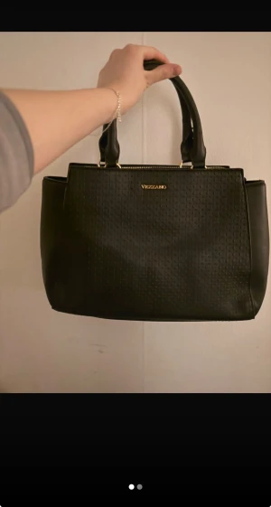 Svart handväska från Vezzano - Stilren svart handväska från Vezzano med guldfärgade detaljer och präglat mönster på framsidan. Väskan har två rejäla handtag, dragkedja upptill och en praktisk ficka med dragkedja på baksidan. Perfekt storlek för dina essentials.
