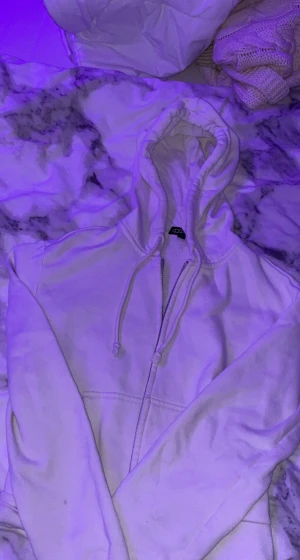 Vit hoodie med dragkedja från H&M - Vit hoodie från H&M med dragkedja framtill och snörning i huvan. Tröjan har långa ärmar och ribbade muddar vid ärmslut och nederkant. Perfekt för chill dagar och enkel att matcha med jeans eller mjukisbyxor.