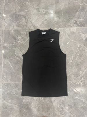 Svart linnetopp från Gymshark - Svart linnetopp från Gymshark med vitt logotryck på bröstet. Toppen är ärmlös och har rund halsringning. Materialet är lätt och andas, perfekt för intensiva träningspass. Snygg och sportig design som ger dig fri rörlighet på gymmet.