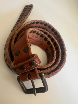Läderskärp från Massimo Dutti - Brunt nitat bälte i skinn från Massimo Dutti med en otroligt mjuk kvalitet och känsla. Skärpet har råa kanter och en robust metallspänne i mörk matt finish. Perfekt till jeans eller som accessoar över tröja. Modellen på bilden har strl 36, passar S/M. 
