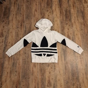 Vit hoodie med svart Adidas-tryck - Säljer en vit hoodie från Adidas med stort svart Trefoil-tryck och svarta detaljer på ärmarna. Tröjan har huva, långa ärmar och ribbade muddar. Materialet är mjuk bomull, perfekt för chill dagar. Adidas-logga även på ena ärmen för extra streetkänsla.
