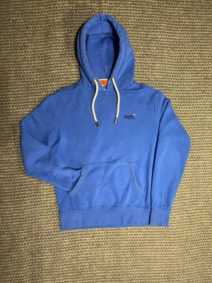 Superdry Hoodie - Säljer en blå hoodie från Superdry Orange Label med vit snörning i huvan och broderad logga på bröstet. Klassisk känguruficka framtill och ribbade muddar. Perfekt för dig som gillar en enkel och stilren look. Om du undrar något är det bara att skriva🙌✅