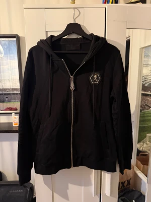 Svart hoodie med orm och döskalle - Svart hoodie från Philipp Plein med dragkedja, huva och snörning. Framsidan har en döskallepatch på bröstet och baksidan pryds av en stor orm i guld och silver. Ärmarna har ribbade detaljer för extra edge. Perfekt för dig som gillar streetstyle.