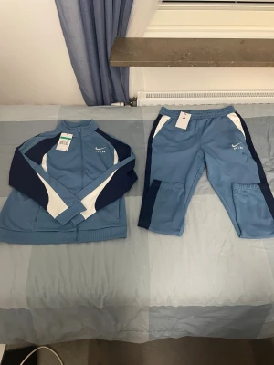 Nike Air blå träningsbyxor XL - Nike Air träningsbyxor i ljusblå med mörkblå och vita paneler. Byxorna har elastisk midja och är gjorda i mjuk polyester, perfekt för sport och chill. Snyggt broderad Nike Air-logga framtill. Matcha med jackan för en komplett look.