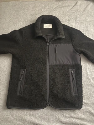 Mörkblå teddyjacka från A Day's March - Mörkblå teddyjacka från A Day's March med mjuk fleeceyta och kontrasterande paneler i syntetmaterial. Jackan har hög krage, tre fickor med dragkedja och hel dragkedja framtill. Perfekt för kalla dagar och har en clean, modern look.