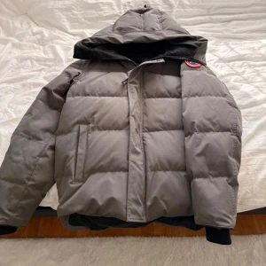 Grå dunjacka från Canada Goose - Säljer en grå dunjacka från Canada Goose med huva och klassisk patch på axeln. Jackan är quiltad, har dragkedja och två sidofickor. Perfekt för kalla dagar och har en clean, stilren look som funkar till det mesta. Finns små fläckar på jackan därför är priset ej hugget i sten 