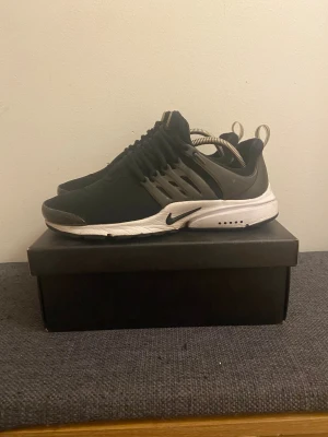Nike Presto svarta sneakers - Nike Presto sneakers i svart med vit sula och grå detaljer. Skorna har en rund tå, snörning och en flexibel syntetisk ovandel som ger en skön passform. Perfekt för dig som gillar sportig stil och vill ha bekväma sneakers med cool design. Litet hål vid höger tå