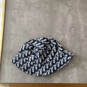 Dior Bucket hat - Snygg bucket hat från Off Work med svart och vit grafiskt mönster. Hatten har klassisk form med nedvikt brätte och är tillverkad i ett stadigt tyg som ger en cool streetkänsla. Perfekt accessoar för att lyfta din outfit.