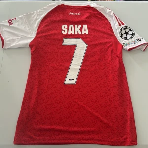 Arsenal Saka 7 Champions League tröja - Arsenal matchtröja med Saka 7-tryck, Champions League-märke på ärmen och Emirates Fly Better-logga. Röd bas med vita ärmar och röda Adidas-ränder, diskret A-mönster över hela tröjan. Tillverkad i lätt och ventilerande polyester. Replika, endast handtvätt gäller!