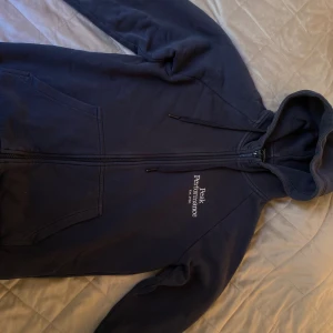 Mörkblå hoodie från Peak Performance Storlek S - Säljer en mörkblå hoodie från Peak Performance med dragkedja och huva. Tröjan har broderad logga på bröstet, kängurufickor och är tillverkad i mjuk bomullsmix. Perfekt för chill dagar eller när du vill ha en sportig look.