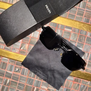 Svarta solglasögon från Prada - Snygga svarta solglasögon från Prada med rektangulära linser och coola detaljer på skalmarna där Prada-loggan syns tydligt. Glasögonen har näskuddar i metall och kommer med originalfodral och putsduk.