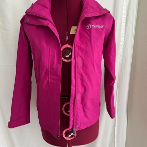Rosa regnjacka från Tenson - Snygg och färgstark rosa vindjacka från Tenson med huva och dragkedja framtill. Jackan har långa ärmar med kardborreband i ärmslut. En jacka för regniga och blåsiga dagar. Fint skick. 