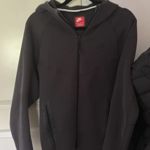 Svart Nike hoodie med dragkedja XS - Svart hoodie från Nike i storlek XS med hel dragkedja framtill och två fickor med dragkedja. Tröjan har huva och är långärmad, perfekt för chill eller träning. Enkel och stilren design med diskret Nike-logga på bröstet.